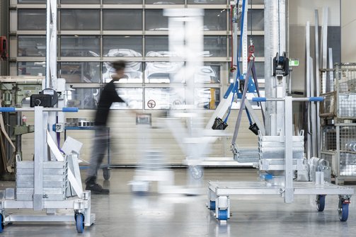 SCHILLING Produktion Ein Mann schiebt eine Last auf Rollen durch die Produktion von SCHILLING in Friedrichshafen, im Hintergrund Metallgestelle und großen Fenstern, die Bewegung ist leicht verschwommen dargestellt.