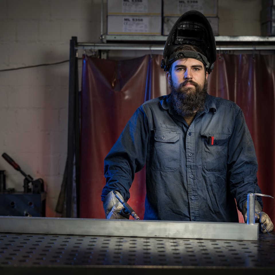 Schweißarbeiten in der Produktion Ein Mann mit Vollbart, Schweißmaske und Arbeitskleidung steht konzentriert an einer Metallschweißstation bei SCHILLING Kran- und Hebetchnik.