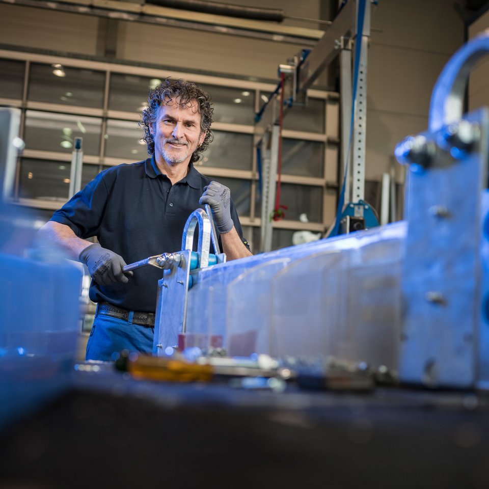 Ein Mann arbeitet in der SCHILLING-Produktion an einem Bauteil aus Aluminium, im Hintergrund sind Regale mit weiteren Materialien und Werkzeugen zu sehen.