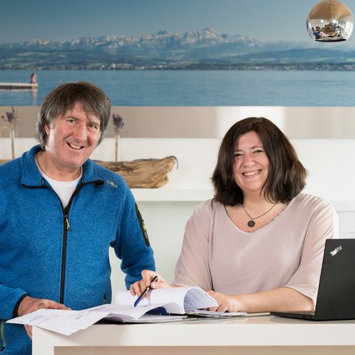 Martin und Heike Schilling Martin und Heike Schilling stehen nebeneinander, lächeln und schauen in die Kamera, vor ihnen geöffnete Unterlagen, daneben steht ein Laptop.