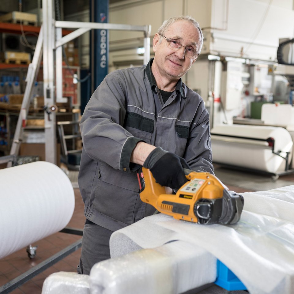 Verpackungsarbeiten Ein Mann in Arbeitskleidung benutzt in der SCHILLING-Produktion eine elektrische Abwickelmaschine, um Verpackungsmaterial um ein Bauteil zu wickeln.