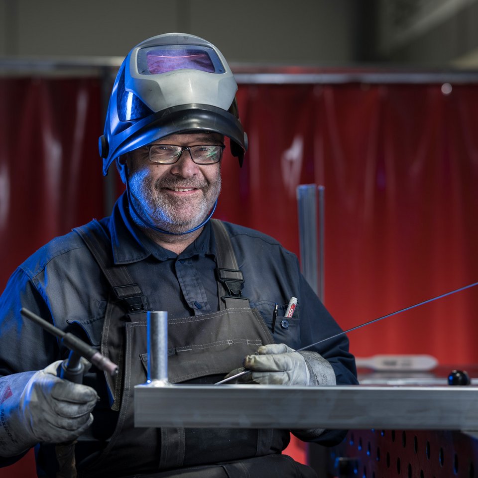 Spaß bei der Arbeit Ein lächelnder Schweißer trägt Schutzhelm und Handschuhe und arbeitet an einem Metalltisch mit Schweißgerät.